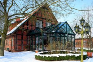 Dieser DecoArt-Wintergarten in Nortrup (Objekt 827) besitzt ein Pultdach mit beidseitigem Walm und eingeschifftem Satteldach. Winterlicher Blick auf ein Fachwerkhaus im Erholungsgebiet Hasetal mit gläsernem Wintergarten-Anbau und verschneitem Gartenbereich.